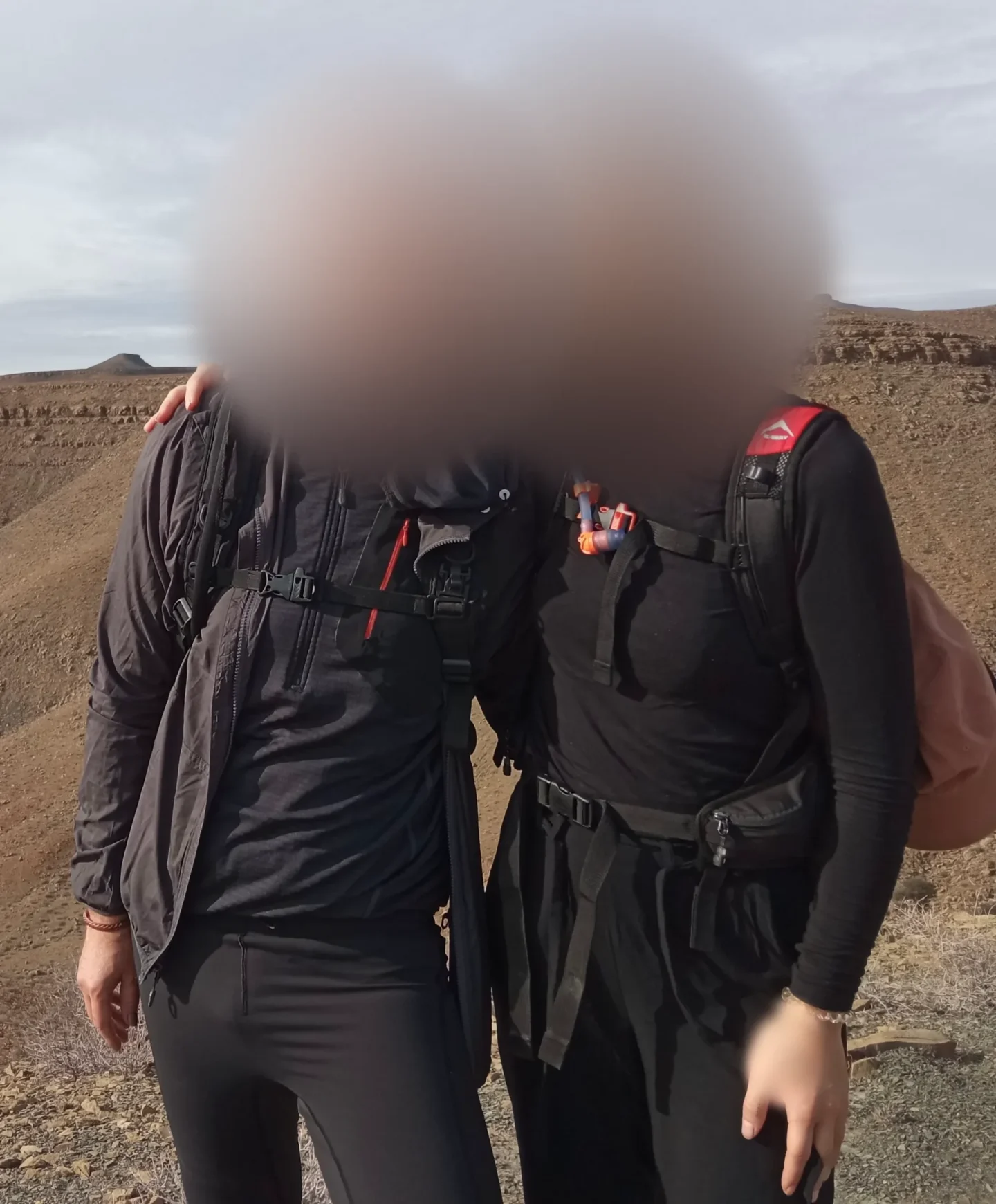 Two hikers in dark outdoor clothing pose arm in arm on a rocky ridge, their faces softly blurred