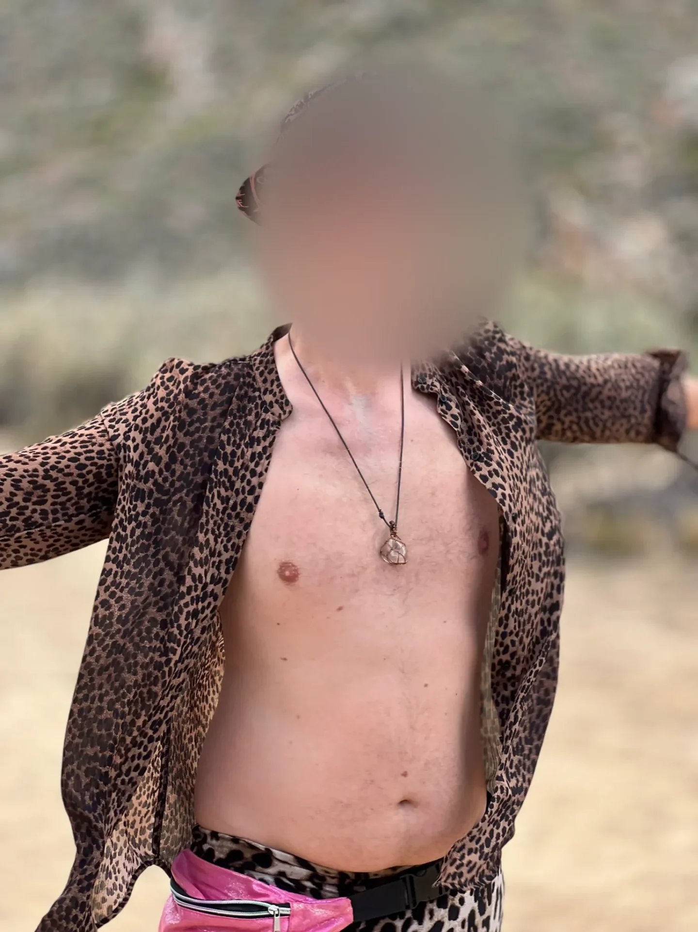The same man from the front in the open leopard-print shirt, a crystal pendant on his chest, arms spread, his face softly blurred