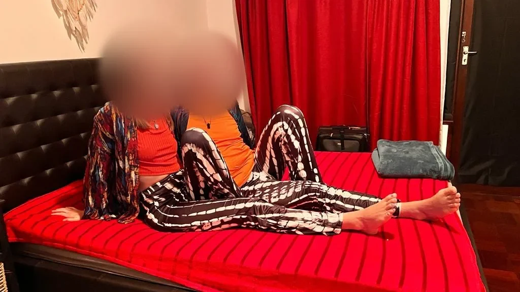 Two smiling people sit on a bed with red covers, both in black-and-white patterned trousers, their faces softly blurred