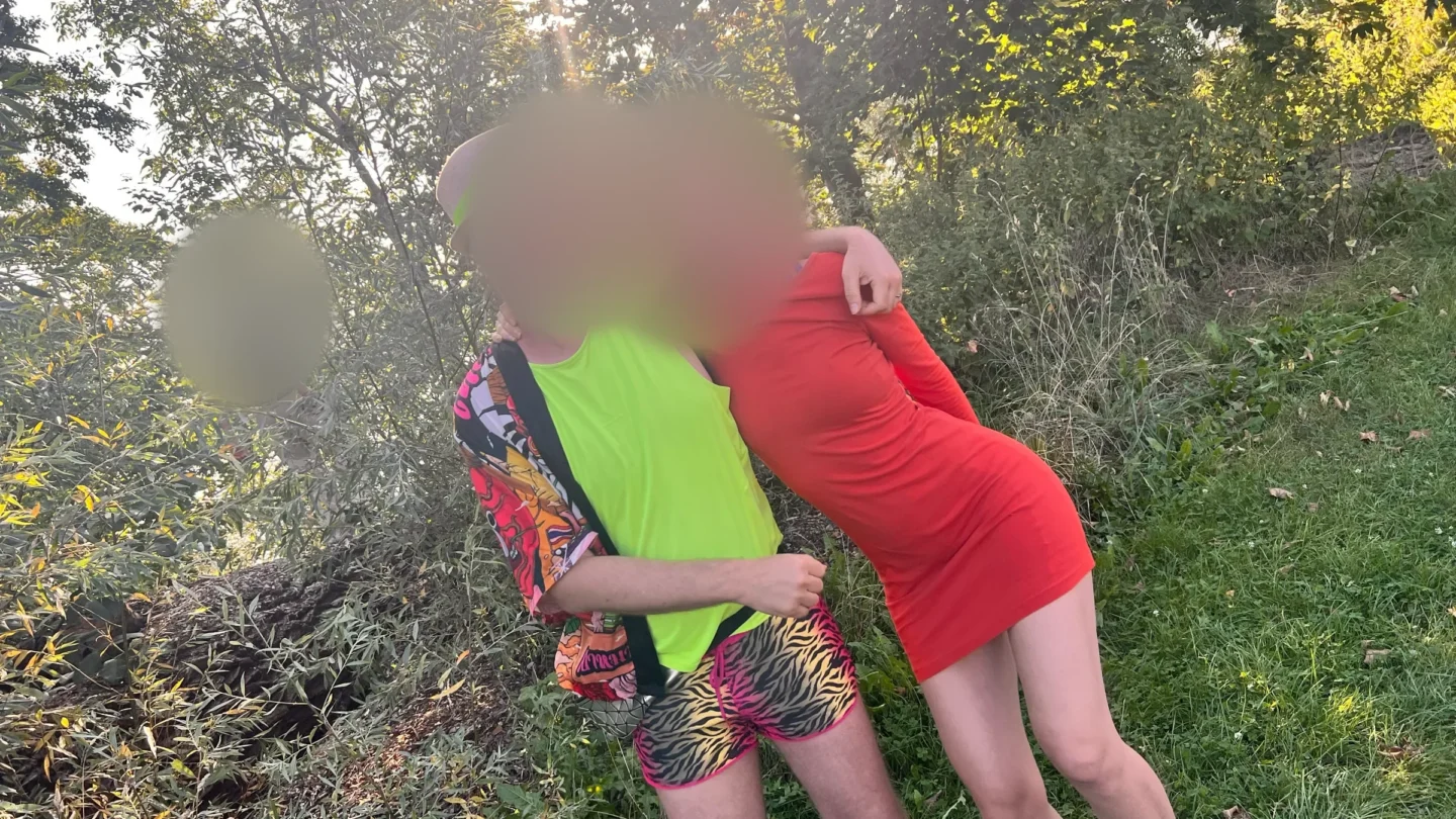 Two people in the golden evening light in a green wooded spot, one in a neon-green top and zebra-print shorts, one in a red dress, their faces softly blurred