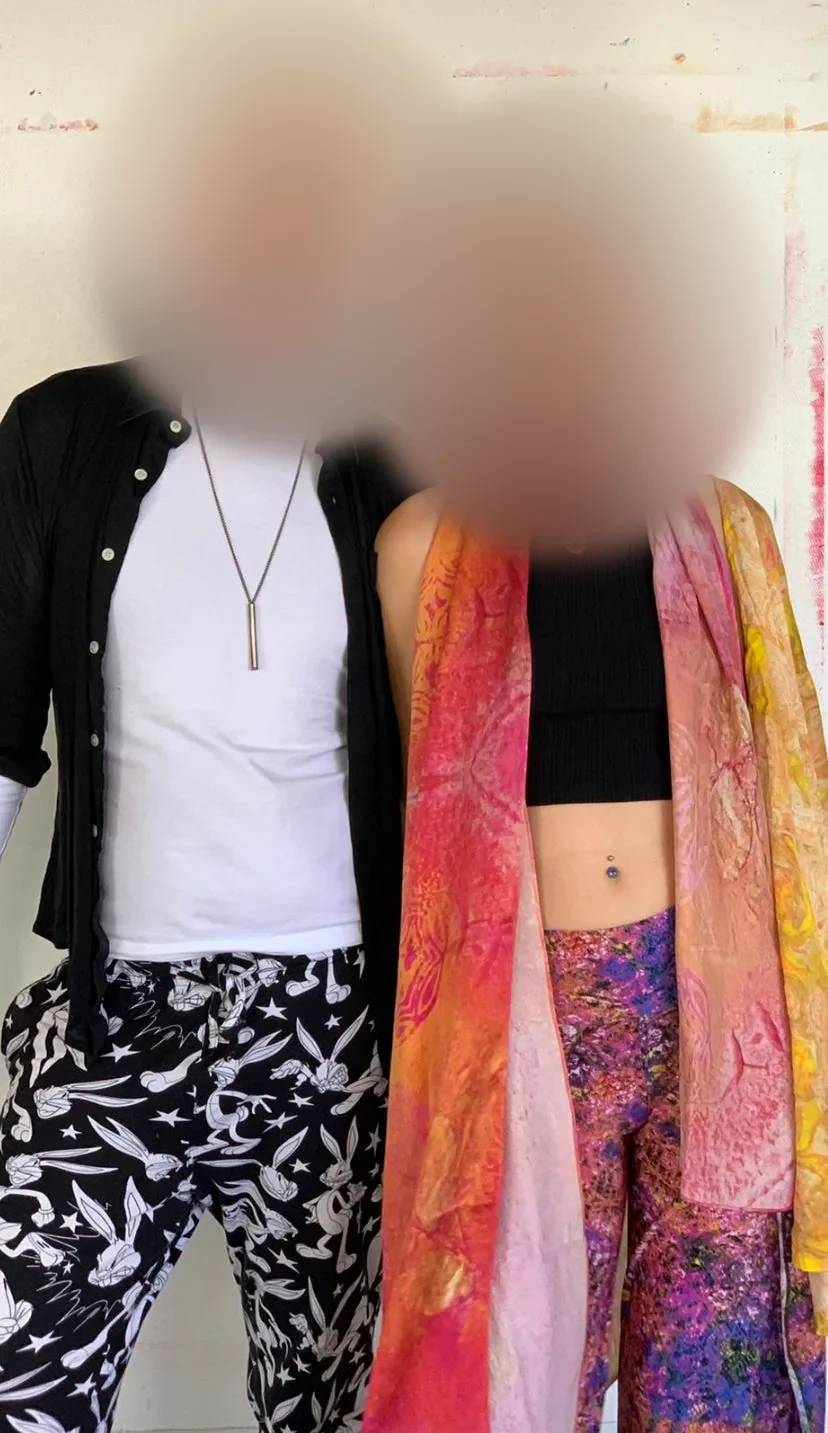 Two people in front of a white wall, one in a black shirt and rabbit-print trousers, one in a flowing rainbow silk scarf, their faces softly blurred