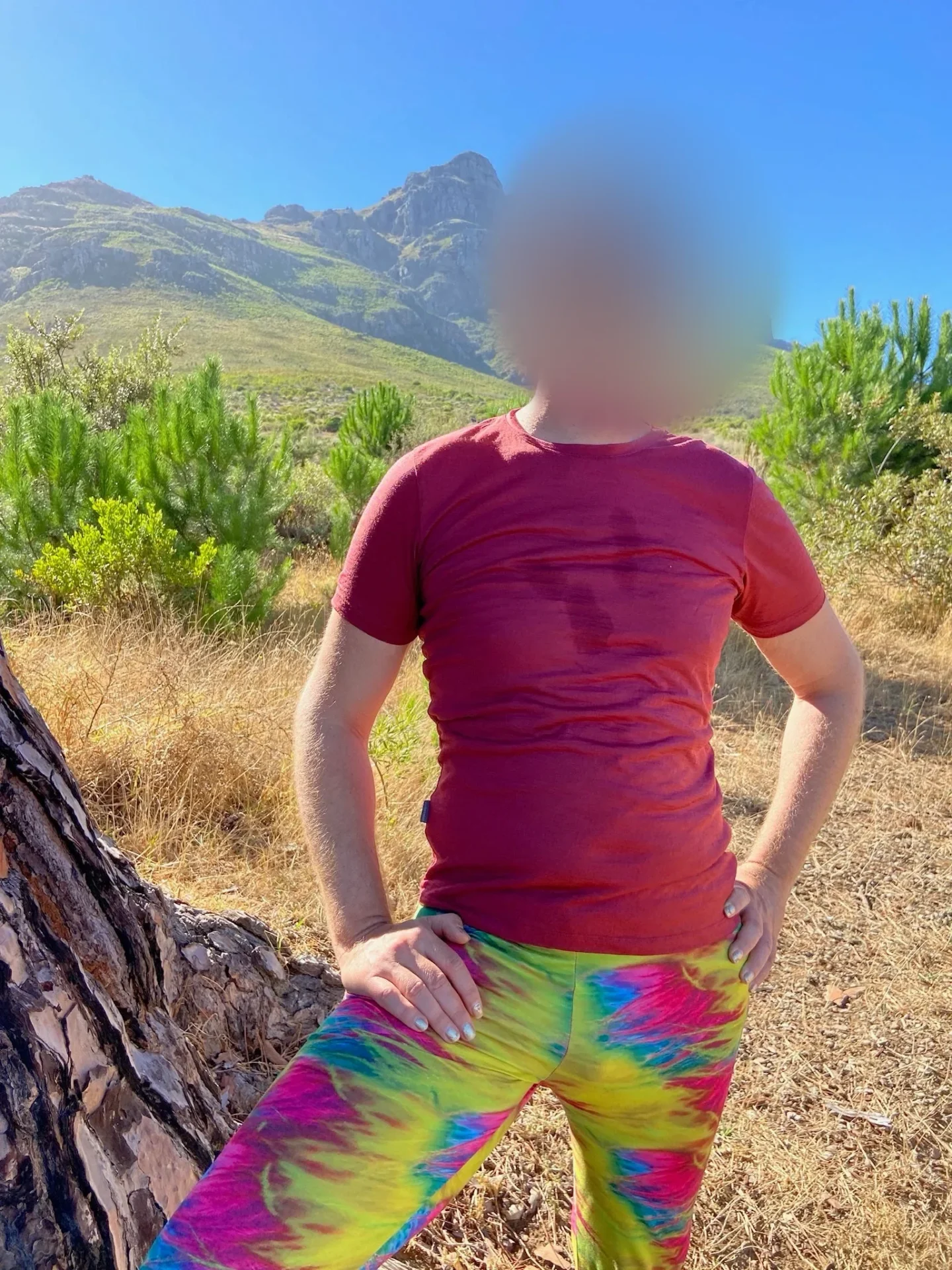 A smiling man in sunglasses, a red t-shirt and tie-dye trousers stands with a hand on his hip in front of a mountain, his face softly blurred