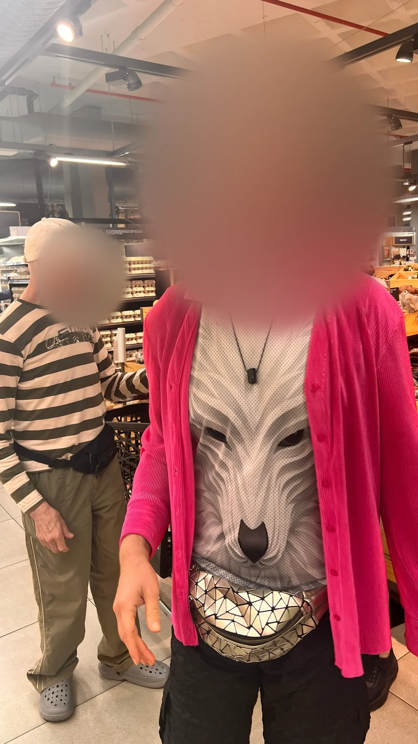 A tall man in a hot-pink shirt over a wolf-print top and a mirrored belt bag stands in a supermarket aisle, his face softly blurred