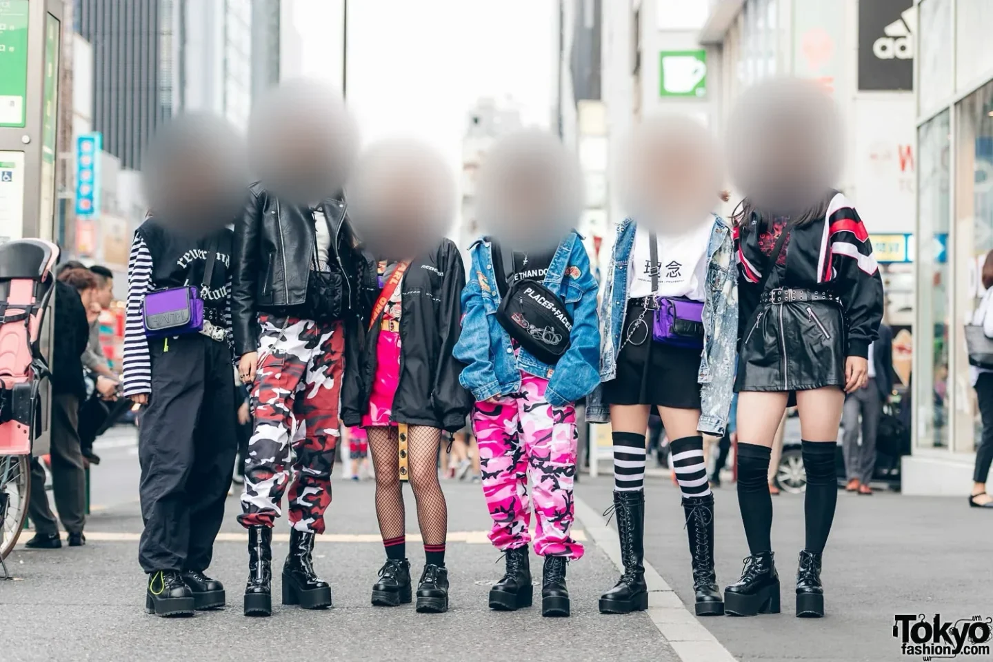 Six young people in layered black, camouflage, denim and pink street outfits stand in a row on a city street, their faces softly blurred