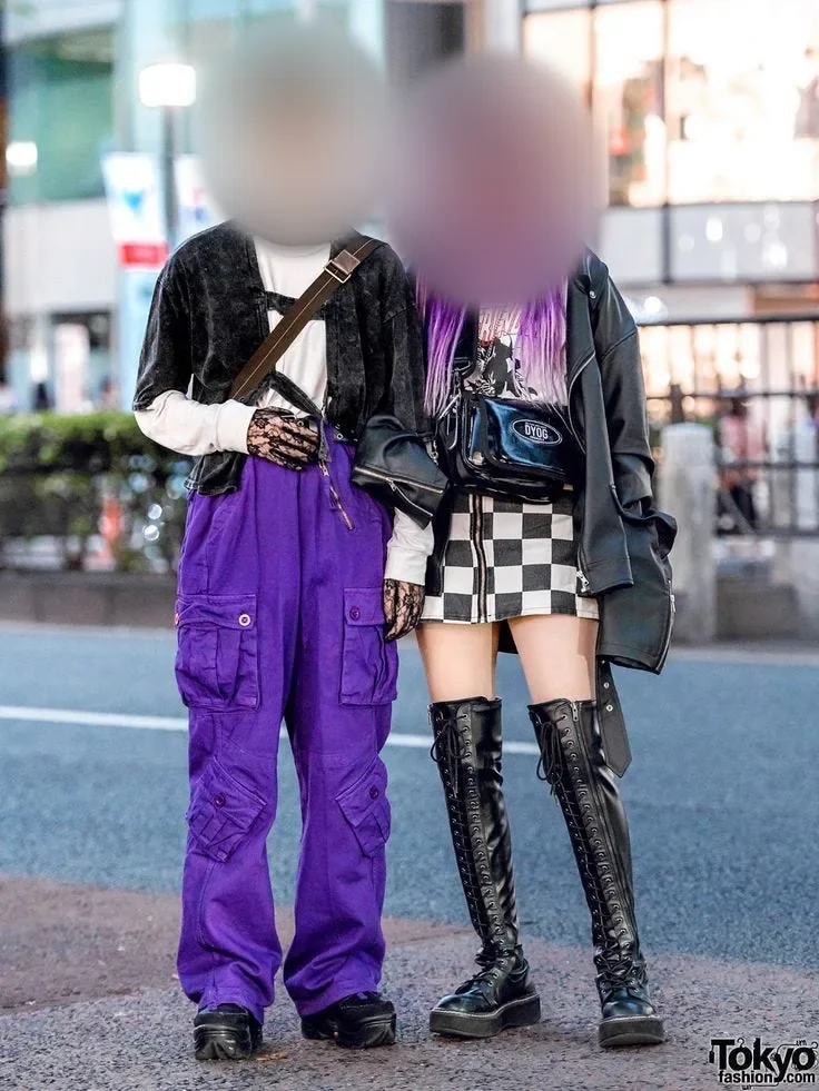 Two young people on a city street: one in a black cardigan and purple cargo trousers, one with long purple pigtails, a checkered skirt and lace-up boots, their faces softly blurred