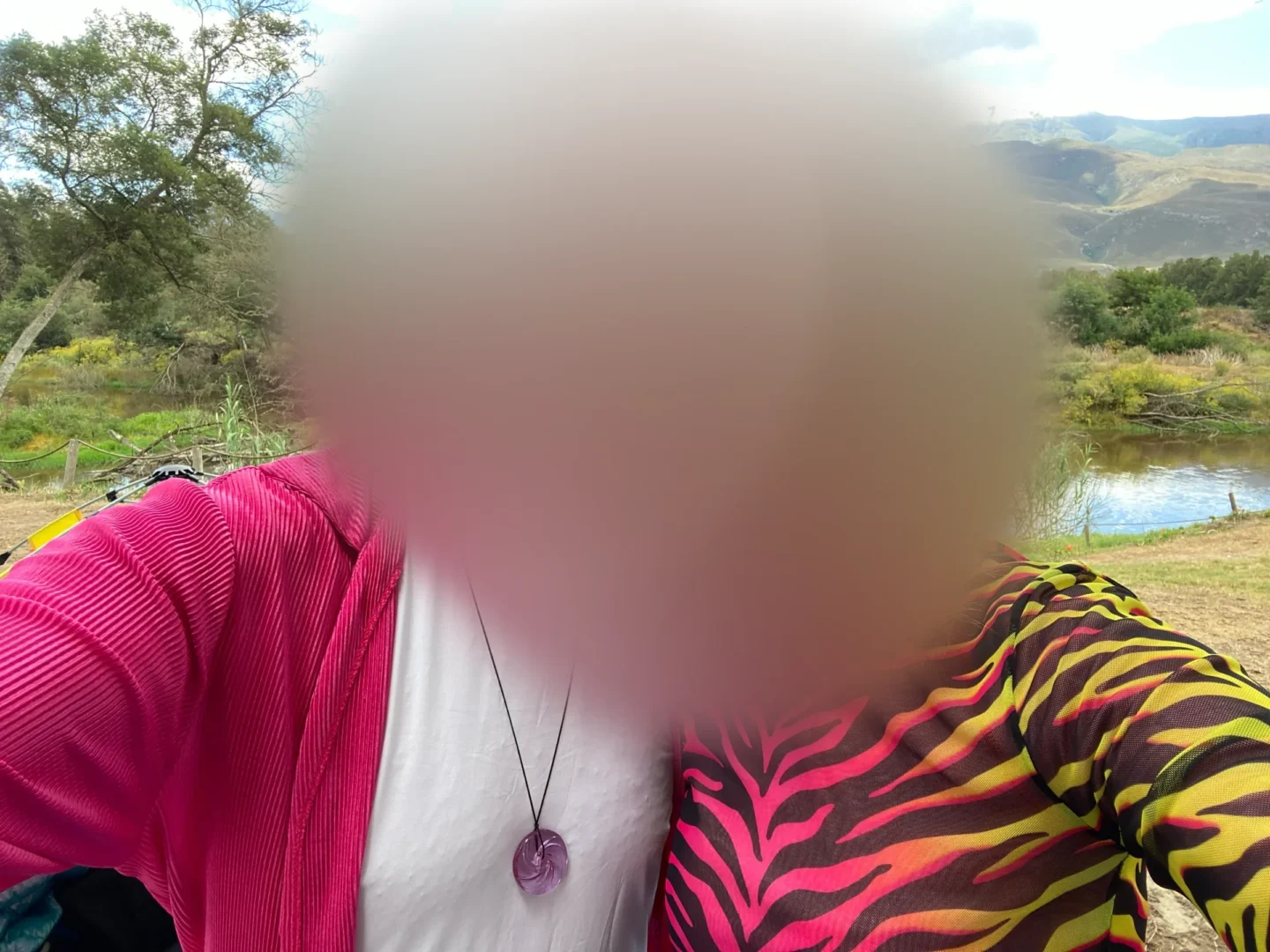 Two people in a selfie by a lake, arms around each other, one in a hot-pink jacket, one in a pink-and-yellow zebra-print top, their faces softly blurred